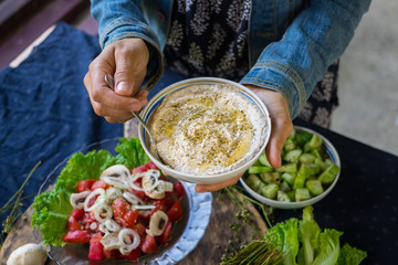 Woman hands adds chickpea hummus spread with sesame seeds tahini to vegetables salad with tomatoes and onion rings on big green salad leaves.  Raw vegan vegetarian food