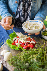Woman hands adds chickpea hummus spread with sesame seeds tahini to vegetables salad with tomatoes and onion rings on big green salad leaves.  Raw vegan vegetarian food