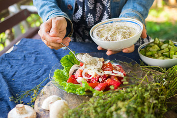 Woman hands adds chickpea hummus spread with sesame seeds tahini to vegetables salad with tomatoes and onion rings on big green salad leaves.  Raw vegan vegetarian food