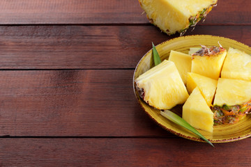 Pieces of pineapple on a yellow plate on wooden boards, pineapple with green leaves on a wooden background, salad of tropical fruits for breakfast, vegetarian food, copy space