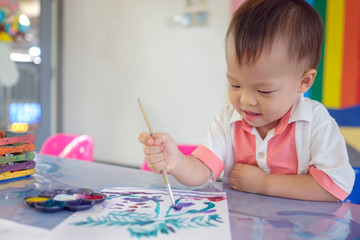 Cute smiling little Asian 18 months / 1 year old toddler baby boy child painting with brush and watercolors at art class, kid painting in child care / play school ,Creative play for toddlers concept
