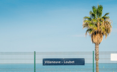 Sign to Villeneuve Loubet with a palm tree and blue Mediterranean Sea, France
