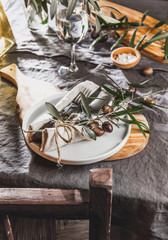 Table setting with gray Linen tablecloth and napkin, white plate, cutlery and and olive tree branch boards decoration. Close up