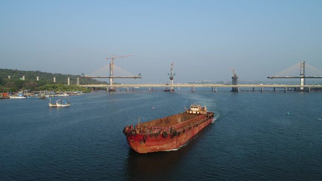 The old rusty and empty barge floats on a large river against the backdrop of a huge bridge under construction. Aerial view.