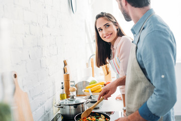 boyfriend frying vegetables on frying pan in kitchen and looking at smiling girlfriend