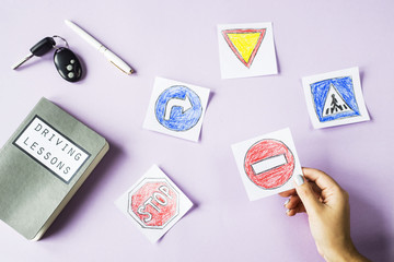 A woman's hand holds a road sign of the stop among other signage drawings next to a workbook for driving lessons. Getting a driving license concept. On a violet background