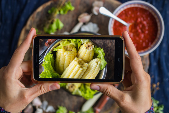 Woman hands take smartphone food photo of cooked corn cobs for lunch or dinner. Phone food photography for social media and blogging. Raw vegan vegetarian