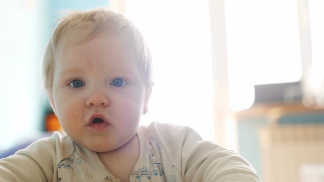 Portrait of healthy serious conscious pretty adorable newborn baby boy standing near sofa and looking at camera bright day light on background chubby cheeks reaching arms blue eyes sweet infant home