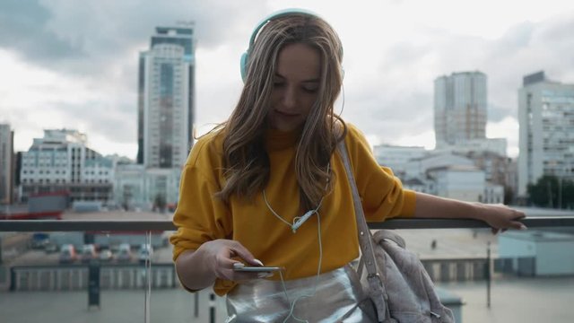 Portrait of young cute attractive young girl in urban city streets background listening to music with headphones. Woman wearing yellow blouse and silver skirt