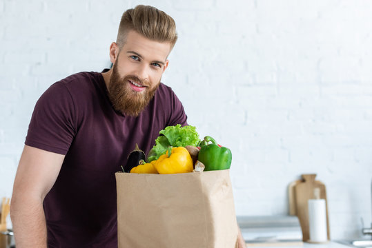 Handsome Bearded Young Man With Grocery Bag Smiling At Camera In Kitchen