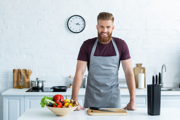 handsome bearded young man in apron smiling at camera while cooking in kitchen - Powered by Adobe