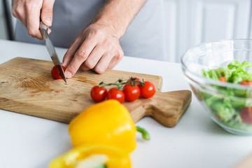 cropped shot of man in apron cutting cherry tomato while cooking salad