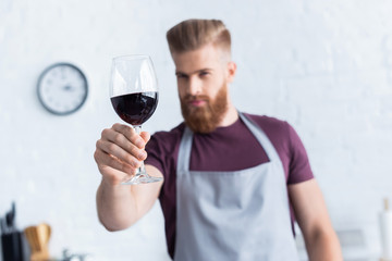 handsome bearded young man in apron holding glass of red wine in kitchen