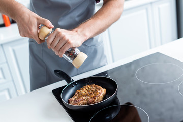 cropped shot of man holding pepper mill while cooking delicious steak in frying pan