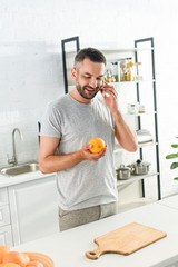 smiling man talking on smartphone and holding orange on kitchen