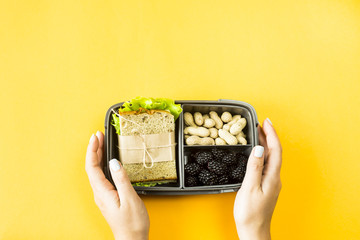 Des mains féminines tiennent une boîte à lunch avec de la nourriture - sandwich, noix et baies sur fond jaune. Vue de dessus, mise à plat,