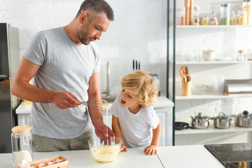 father holding whisk over bowl while his son trying to lick whisk with dough at kitchen