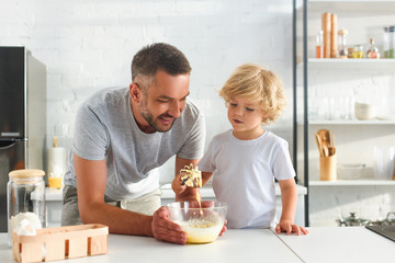 smiling man holding bowl while his son whisking dough at kitchen