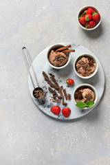 Fototapeta na wymiar Three servings of chocolate ice cream with chocolate, mint and strawberry in white bowls on round stone board. Close-up, top view on gray concrete background