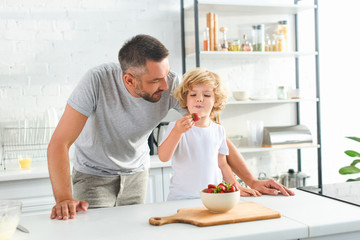 father standing near son while he eating strawberry at kitchen