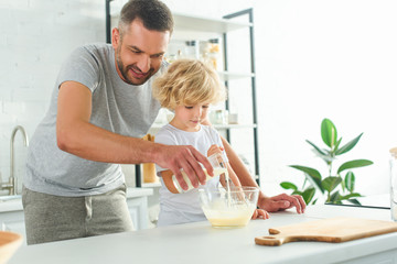 Obraz na płótnie Canvas adorable boy whisking dough while his father pouring milk into bowl at kitchen