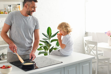 man making pancakes in frying pan while his son sitting near on tabletop at kitchen