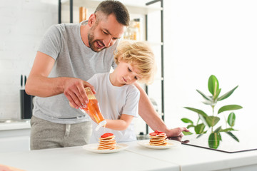 smiling father and son pouring syrup on pancakes at kitchen