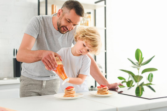 Smiling Father And Son Pouring Syrup On Pancakes At Kitchen