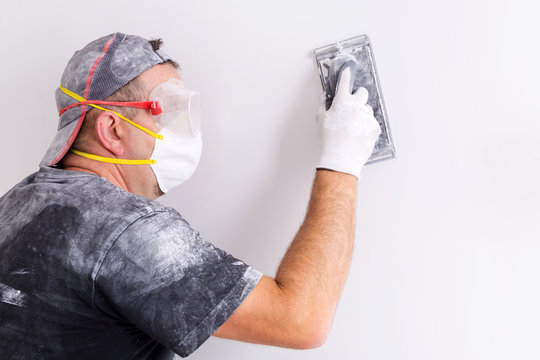 Plasterer wearing dust mask and googles at wall polishing. House renovation concept.