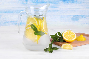 Homemade lemonade with lemon and mint in a glass jug next to a fresh lemon on a white and blue wooden background