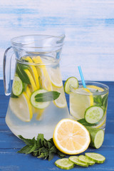Refreshing water with lemon and cucumber and mint in a glass jug and a glass next to fresh lemon on a blue and blue wooden background