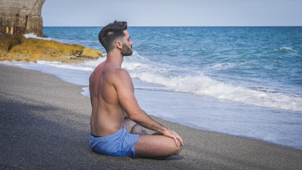 Handsome Shirtless Young Man During Meditation or Doing an Outdoor Yoga Exercise Sitting on Beach Sand by Ocean Shore