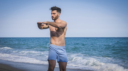 Athletic attractive young man exercising and stretching by the sea, on sandy beach