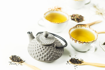 Gray iron kettle among different kinds of dry tea and cups with a ready drink on a white table