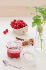 Fresh home made raspberry jam in glass with cinnamon and mint herbs on white wooden table, paper basket with raspberries in background. The light from back.