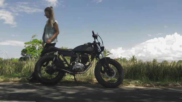 Sexy girl in casual jeans with classic style cafe racer motorcycle. Modern woman try to kick start her old motorcycle. Bike custom made in vintage garage. Brutal fun urban lifestyle. Outdoor portrait.