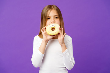 Girl holding big doughnut with icing. Sweet treat, glazed bagel with eyes. Kid eating funny dessert, sweet childhood. Child with long hair wearing white outfit isolated on violet background