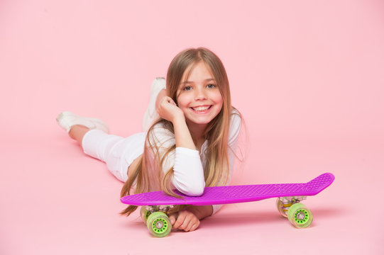 Kid in white outfit lying on floor. Girl with long blond hair isolated on pink background. Child with cute smile leaning on violet skateboard. Teenager having fun, relaxation concept