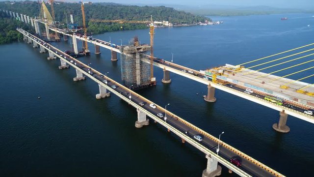 Construction of a new huge cable-stayed bridge over the river. Cars go on the old part of the bridge. Tower cranes over the bridge across the Mandovi river in Goa, India. Shooting with a drone.