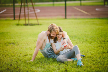 Fototapeta premium Young heterosexual Caucasian man with a beard resting after a workout outdoors in a park on the lawn, green grass sitting in an embrace and using a telphone, watching the screen in red case at sunset