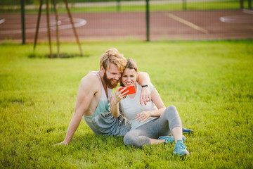 Fototapeta premium Sport and technology. Young in love heterosexual Caucasian couple resting after workout outdoors in park on lawn, green grass sitting in embrace and making selfie photo on smartphone camera at sunset