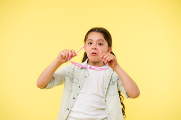 Let me see. Kid girl puts heart shaped eyeglasses. Girl serious face putting glasses on. Child serious holds eyeglasses yellow background. Kid grimace face needs funny eyeglasses