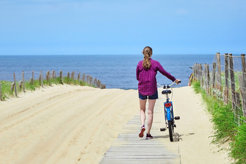 Young woman walking with a bicycle on a wooden path to a sandy beach. Girl looking at the sea or the ocean. Summer sunny day