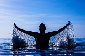 Freeze motion of silhouette of young man splashing water with open arms on water at mystic sunset...