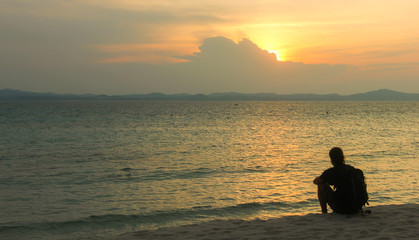 Naklejka na ściany i meble Silhouette of young man with a backpack sitting on the sand staring at sunset on the beach in the island of Kapas, Malaysia. Backpacker enjoying peaceful twilight alone. Summer travel vacation concept