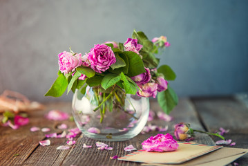 Vintage concept - bouquet of wilting tea roses in glass vase, old letters in craft paper envelopes, blank greeting card on the old wooden rustic background. Retro toned. Selective focus. copy space.