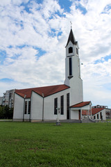 Modern built white church with and black roof located in front of buildings and with large grass area and small stone cross in front on a cloudy day