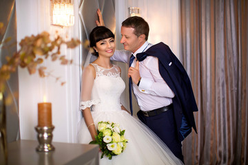 Beautiful bride in white wedding dress is standing near wall, an elegant groom stands nearby and...