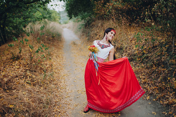 Fototapeta premium Young beautiful girl in Ukrainian national costume with ornament. Handmade decorations. Earrings, necklace. The bride with a bouquet of viburnum is dressed in ethnic style. Long red silk skirt.