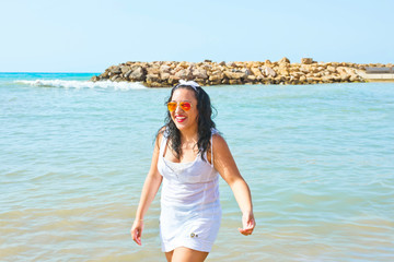 Beautiful Young Brunette Woman with Long Curly Hair in Colorful Sunglasses White Dress Stands in Sea Smiling Laughing. Bright Sunlight Vivid Colors Blue Sky. Summer Vacation Fun Freedom